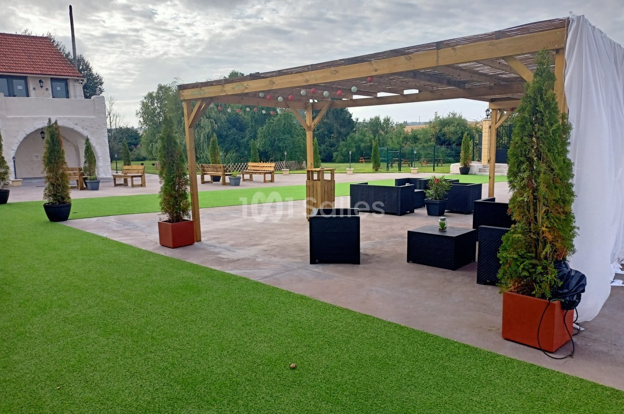 Pergola en bois avec mobilier de jardin noir, entourée de pelouse synthétique et de plantes en pots.
