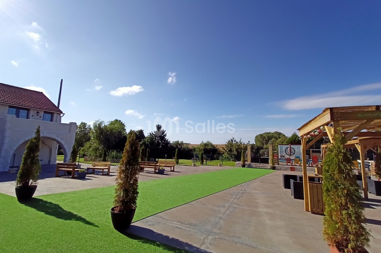 Espace extérieur avec pelouse artificielle, bancs en bois, pergola et ciel dégagé sous une lumière ensoleillée.
