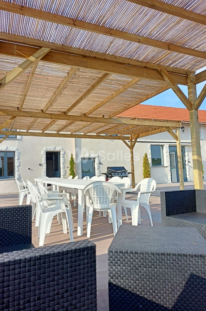 Terrasse avec pergola en bois, table et chaises blanches, entourée de fauteuils en rotin noir devant un bâtiment clair.