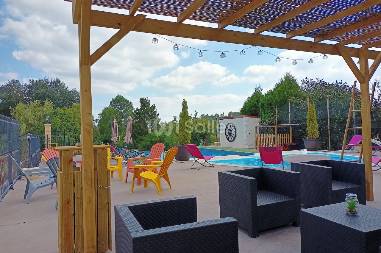 Terrasse avec pergola en bois, mobilier de jardin, piscine et chaises colorées, entourée de verdure sous un ciel…
