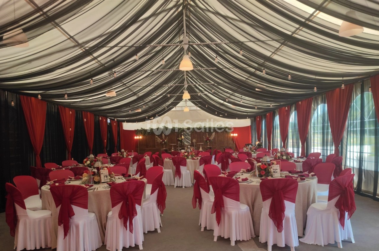 Salle de réception décorée avec des nappes blanches, des chaises ornées de nœuds rouges et des drapés noirs au plafond.