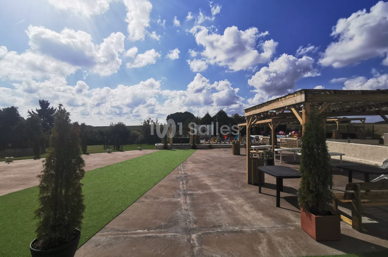 Espace extérieur avec terrasse aménagée, tables, pergola et allées en gazon synthétique sous un ciel partiellement nuageux.
