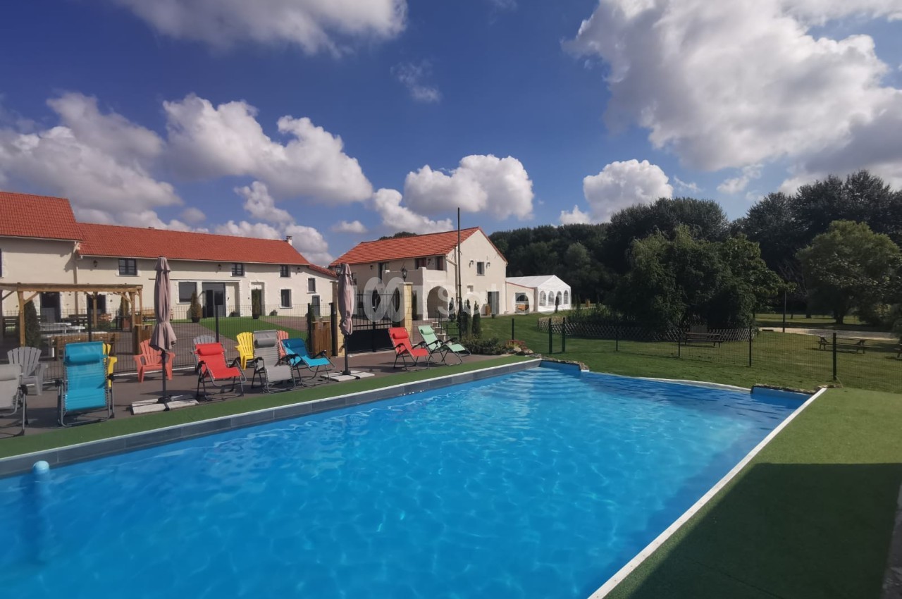 Piscine extérieure avec transats colorés, entourée de bâtiments à toits rouges et d'un jardin arboré sous un ciel bleu.