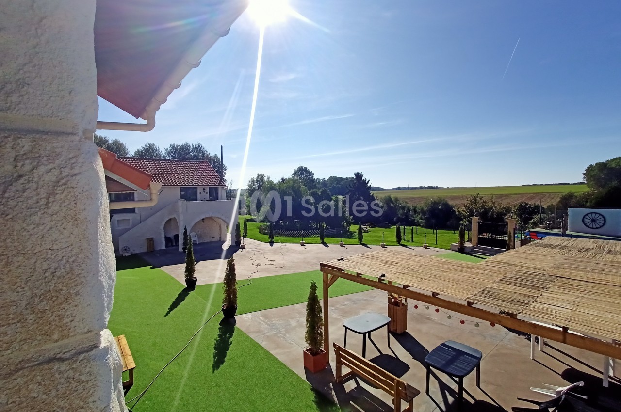 Vue d'une cour ensoleillée avec pelouse, pergola en bois, mobilier de jardin et paysage rural en arrière-plan.