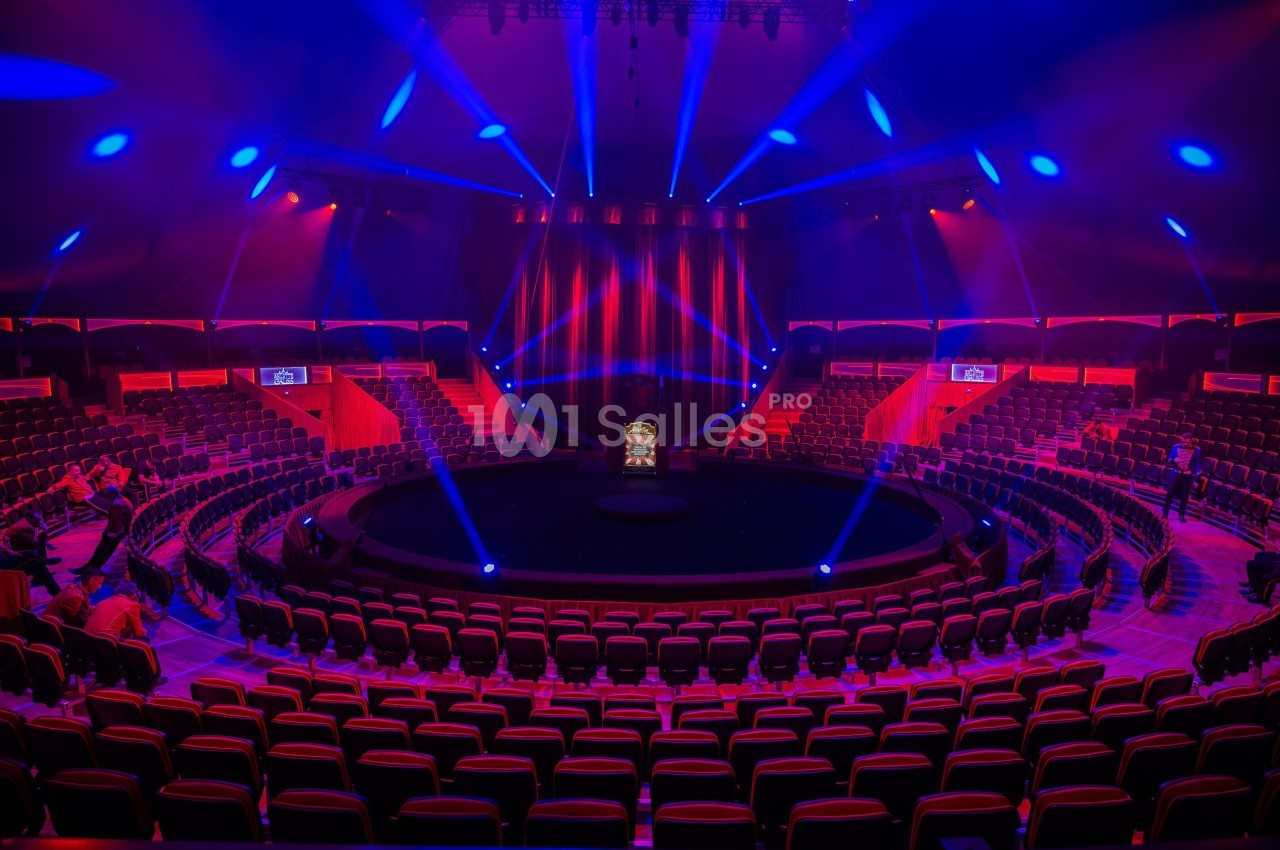 Salle de spectacle circulaire avec sièges rouges, scène centrale éclairée par des projecteurs bleus et rouges.