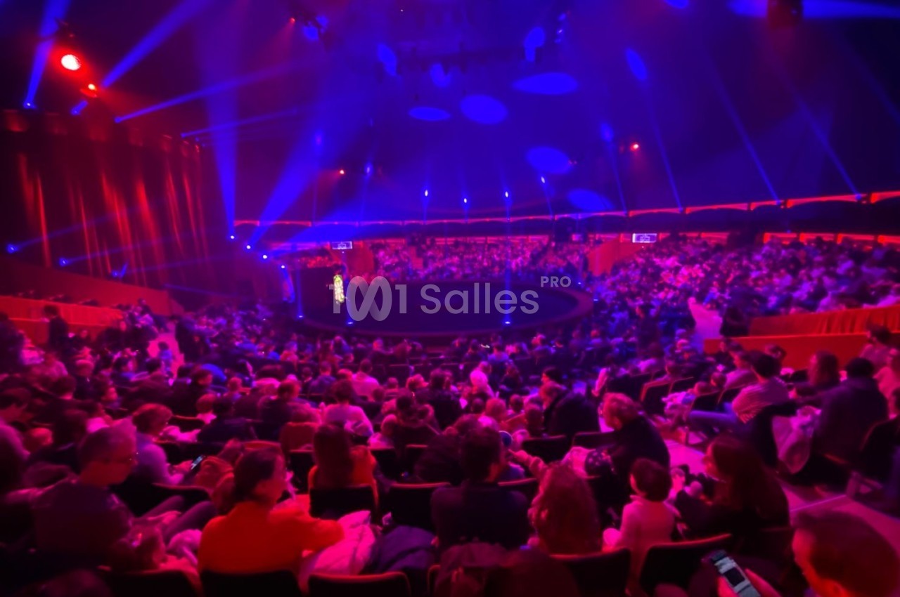 Public assis dans une salle de spectacle circulaire, éclairée par des lumières rouges et bleues.