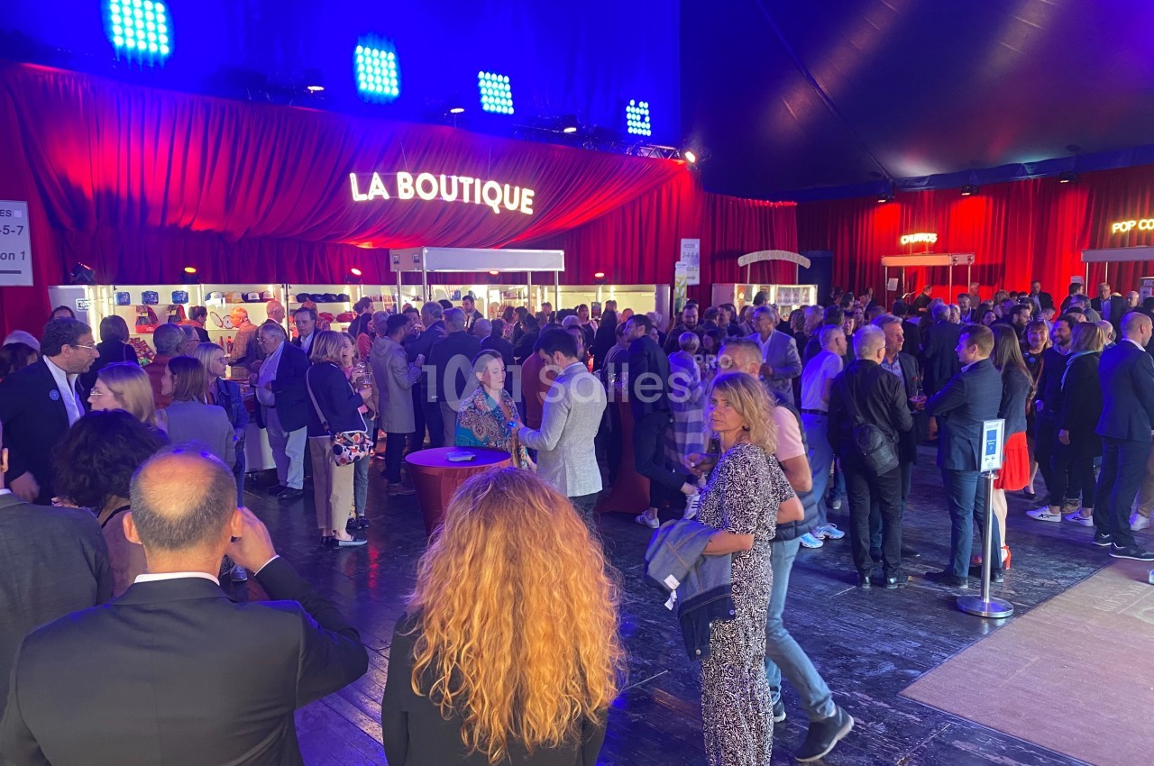 Foule rassemblée dans un espace intérieur éclairé, devant une boutique avec des rideaux rouges en arrière-plan.