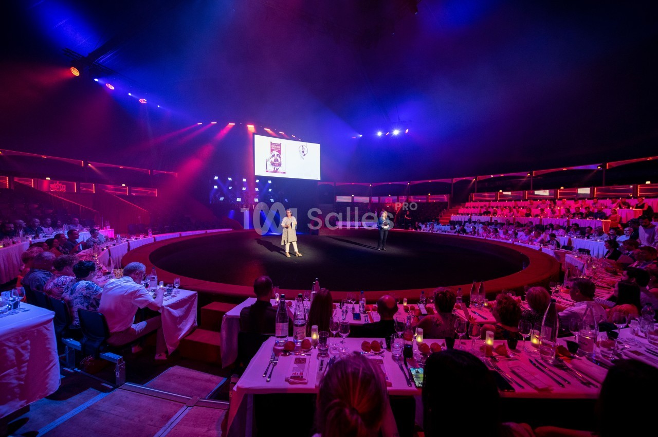Deux personnes sur une scène circulaire éclairée, entourée de spectateurs assis à des tables dans une ambiance tamisée.