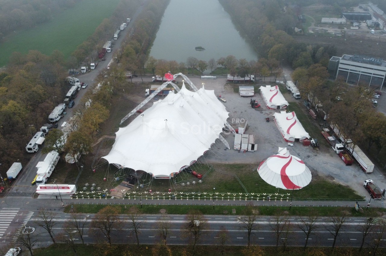 Vue aérienne d'un site de cirque avec plusieurs chapiteaux blancs et rouges installés près d'un canal et d'une route.