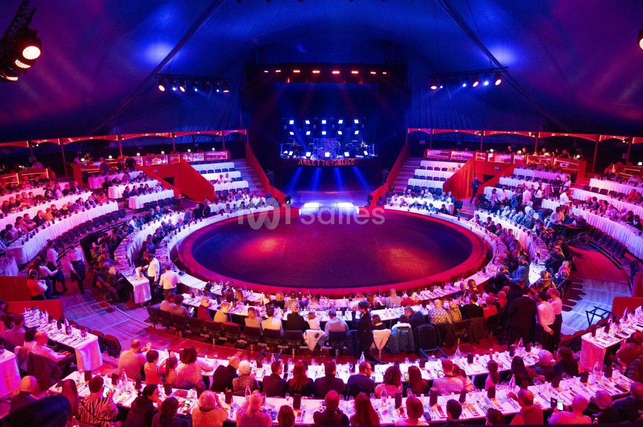 Vue intérieure d'un chapiteau de cirque avec des tables dressées autour d'une piste circulaire et une scène éclairée.