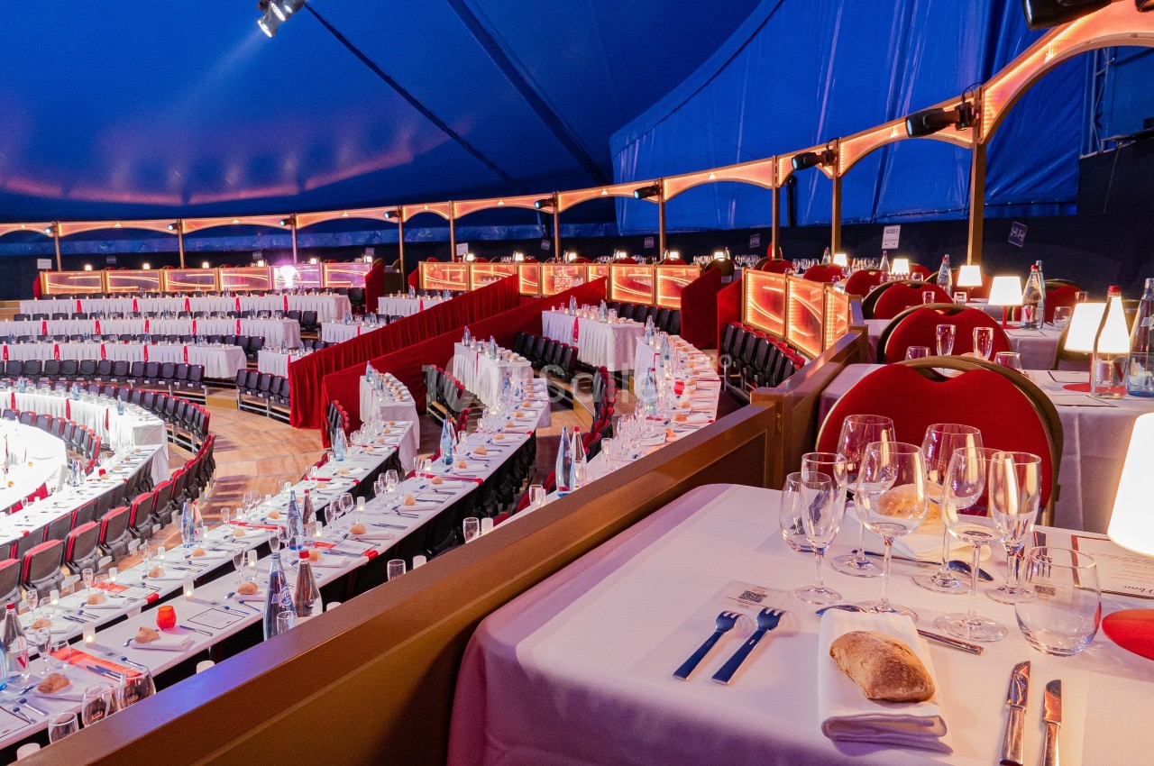 Salle de spectacle sous chapiteau avec tables dressées, nappes blanches et éclairage chaleureux.