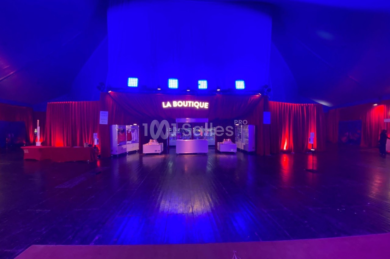 Intérieur d'un chapiteau avec un stand lumineux ’La Boutique’ entouré de rideaux rouges et éclairages bleus.