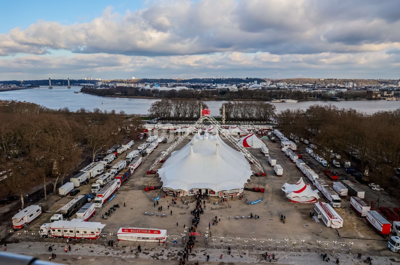 Vue aérienne d'un grand chapiteau blanc entouré de caravanes et de visiteurs, près d'un fleuve et d'une ville.