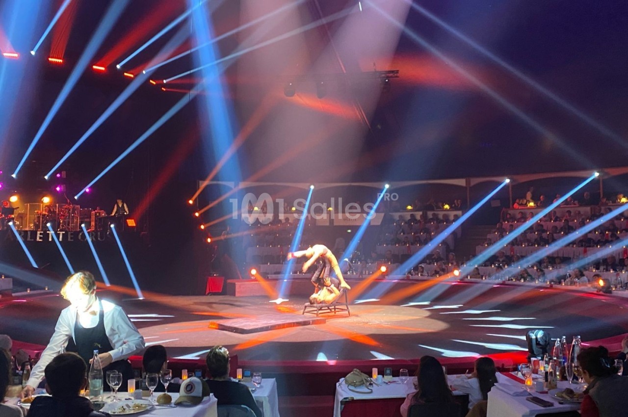 Un artiste réalise une performance acrobatique sur une chaise au centre d'une scène éclairée, devant un public assis.