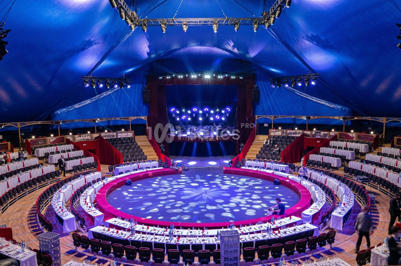 Vue intérieure d'un chapiteau de cirque avec une scène éclairée, des tables dressées et des gradins disposés en cercle.