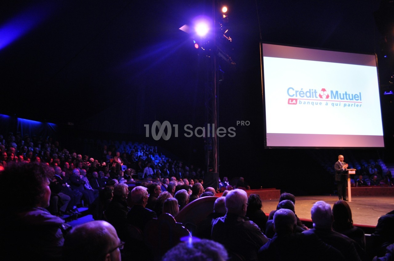 Un conférencier s'adresse à un public nombreux dans une salle sombre avec un écran affichant le logo Crédit Mutuel.