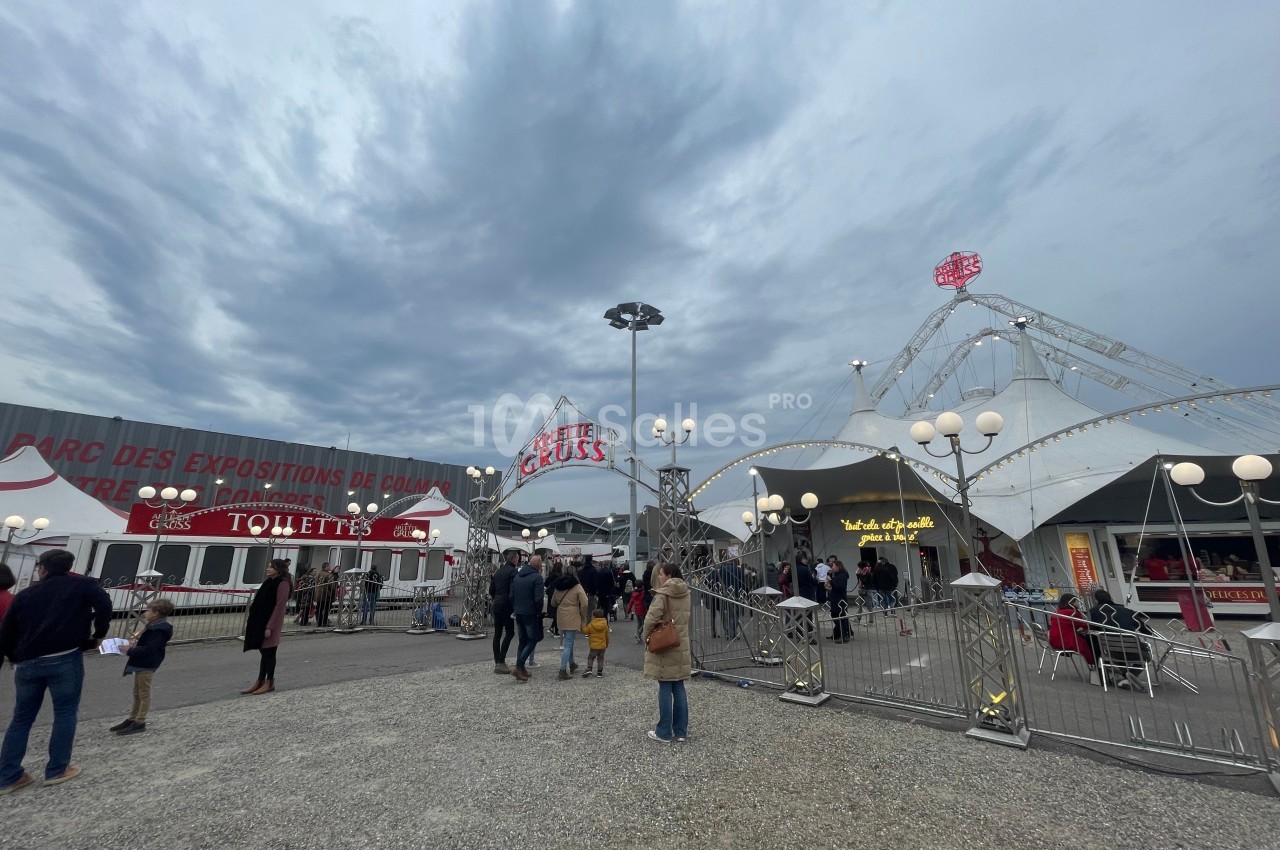 Entrée d'un cirque avec des visiteurs, des tentes blanches, des lampadaires et un ciel nuageux en arrière-plan.