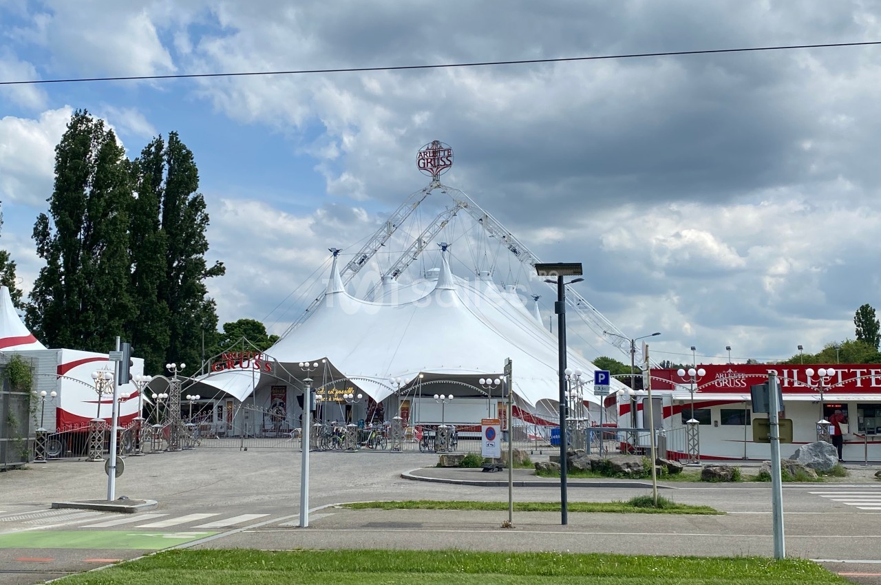 Un grand chapiteau blanc de cirque entouré de stands et de barrières, sous un ciel partiellement nuageux.