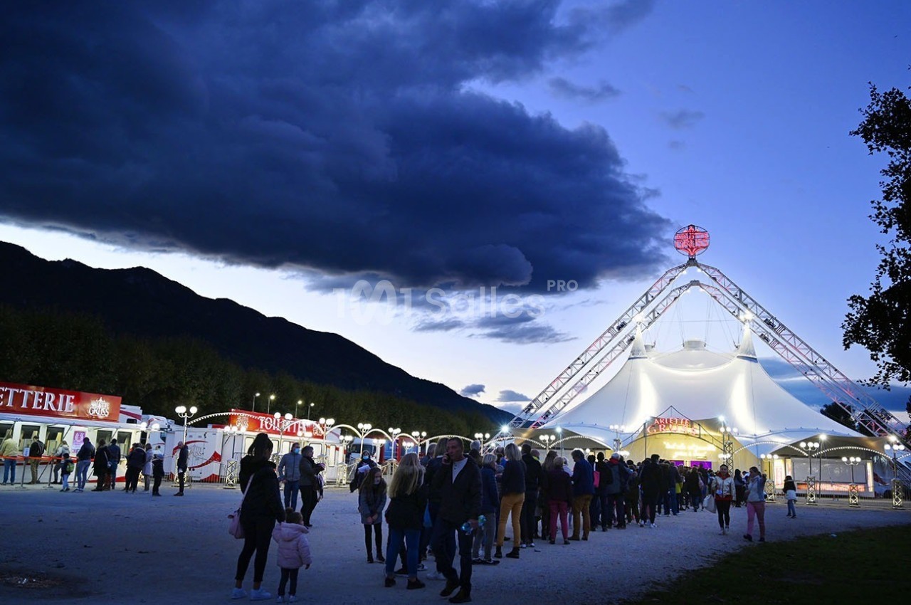 Foule faisant la queue devant un chapiteau éclairé au crépuscule, avec des montagnes et des stands en arrière-plan.