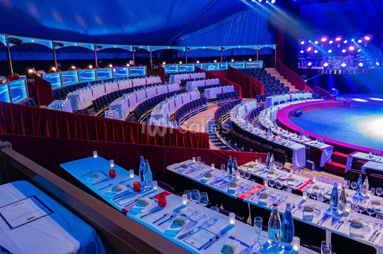 Gradins et tables dressées sous un chapiteau de cirque, éclairés par des lumières colorées.