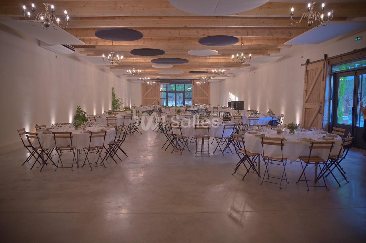 Salle de réception spacieuse avec tables rondes dressées, chaises pliantes en bois et lumière naturelle entrant par de…
