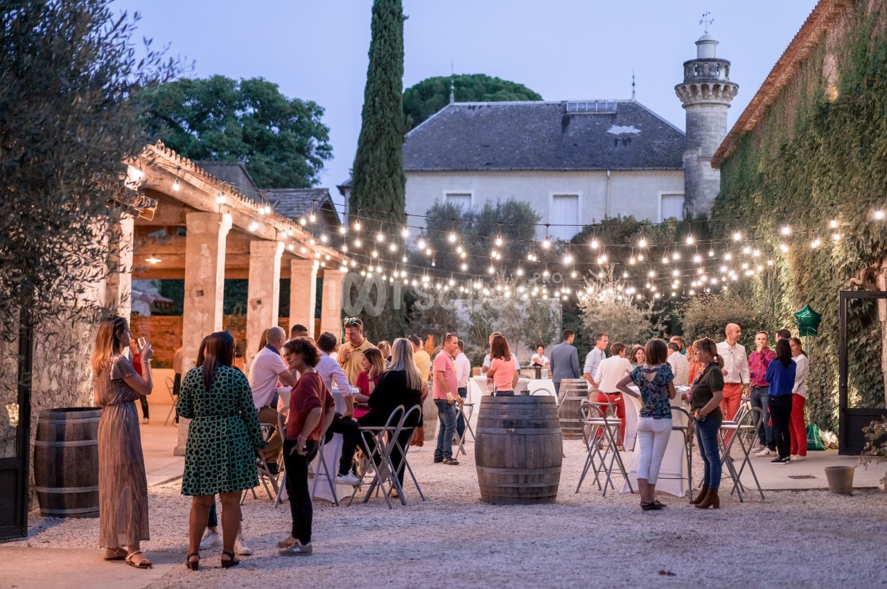 Personnes rassemblées dans une cour extérieure illuminée par des guirlandes, discutant autour de tables et tonneaux.