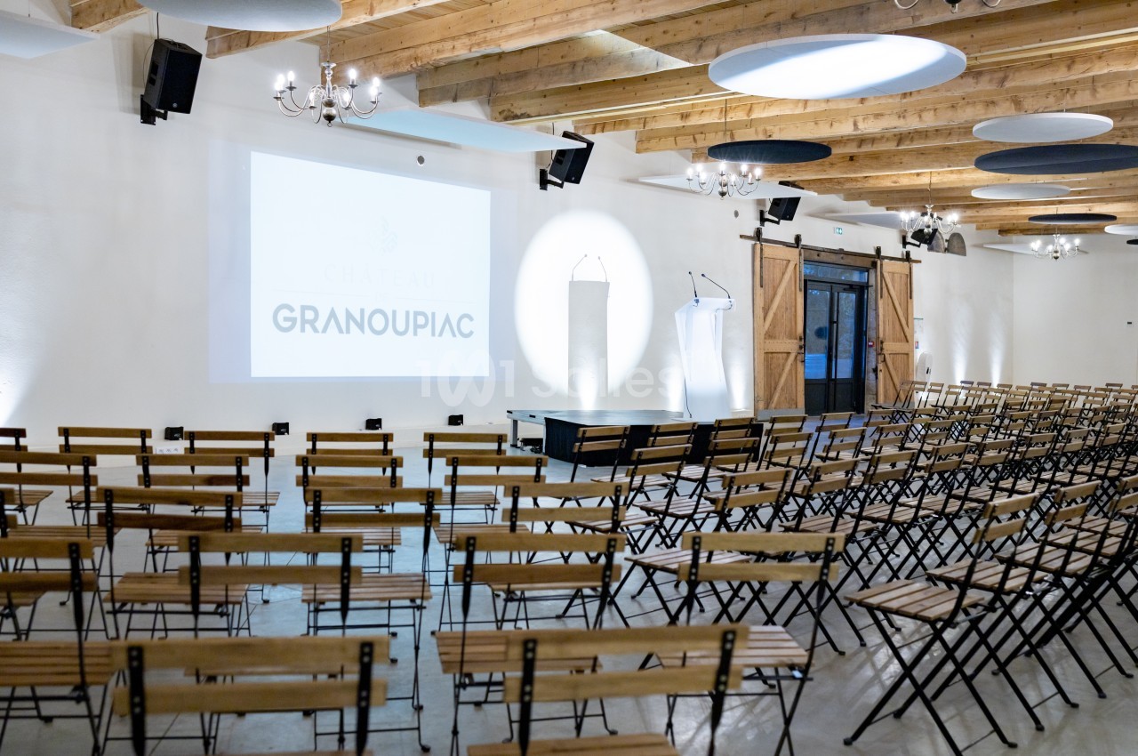 Salle de conférence lumineuse avec des rangées de chaises en bois, un écran de projection et deux pupitres.