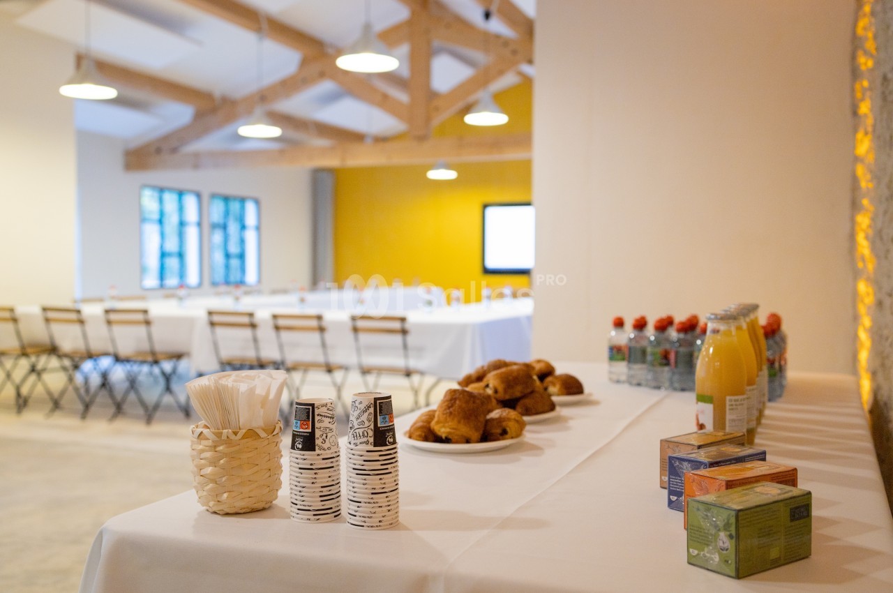 Salle de réunion lumineuse avec tables disposées en U, et buffet comprenant viennoiseries, boissons et gobelets.