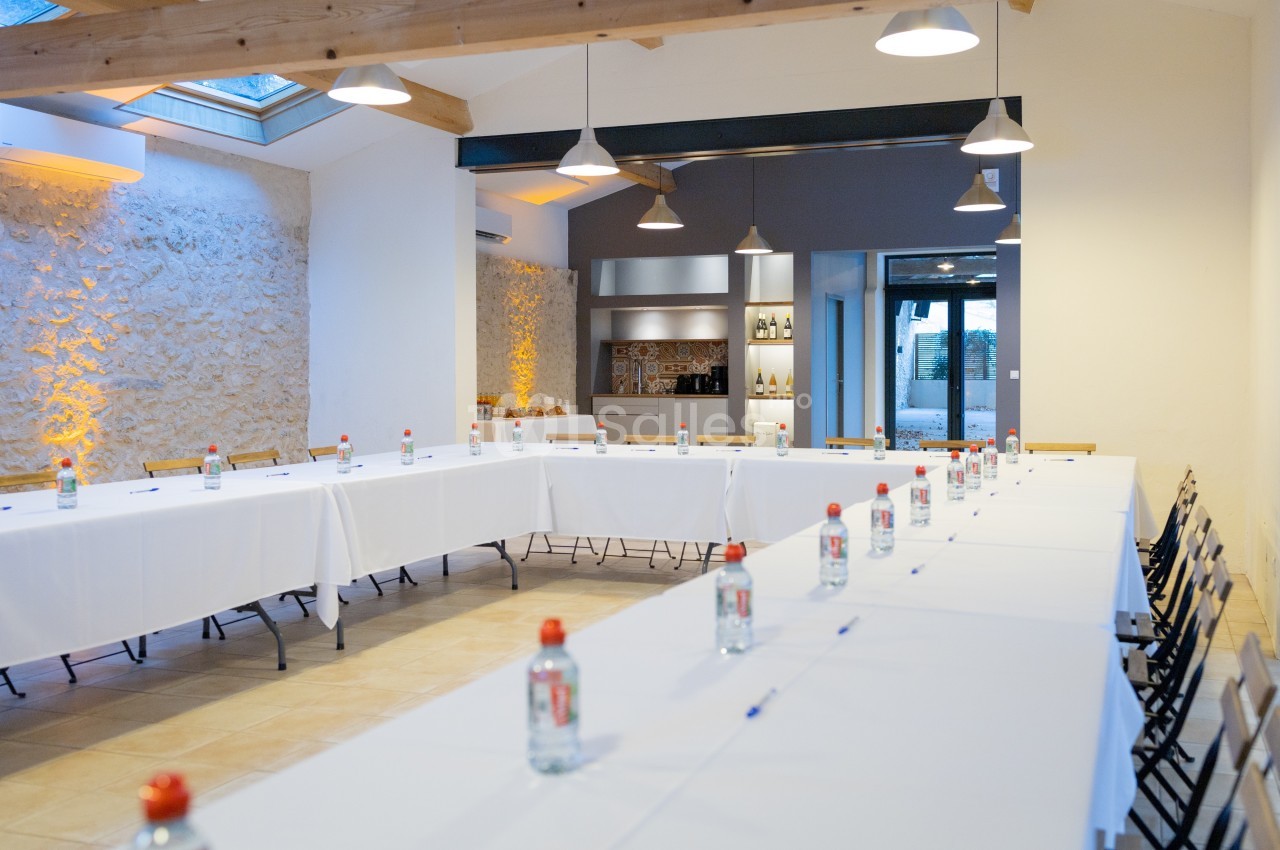 Salle de réunion lumineuse avec tables disposées en U, bouteilles d'eau et stylos, éclairée par des lampes suspendues.
