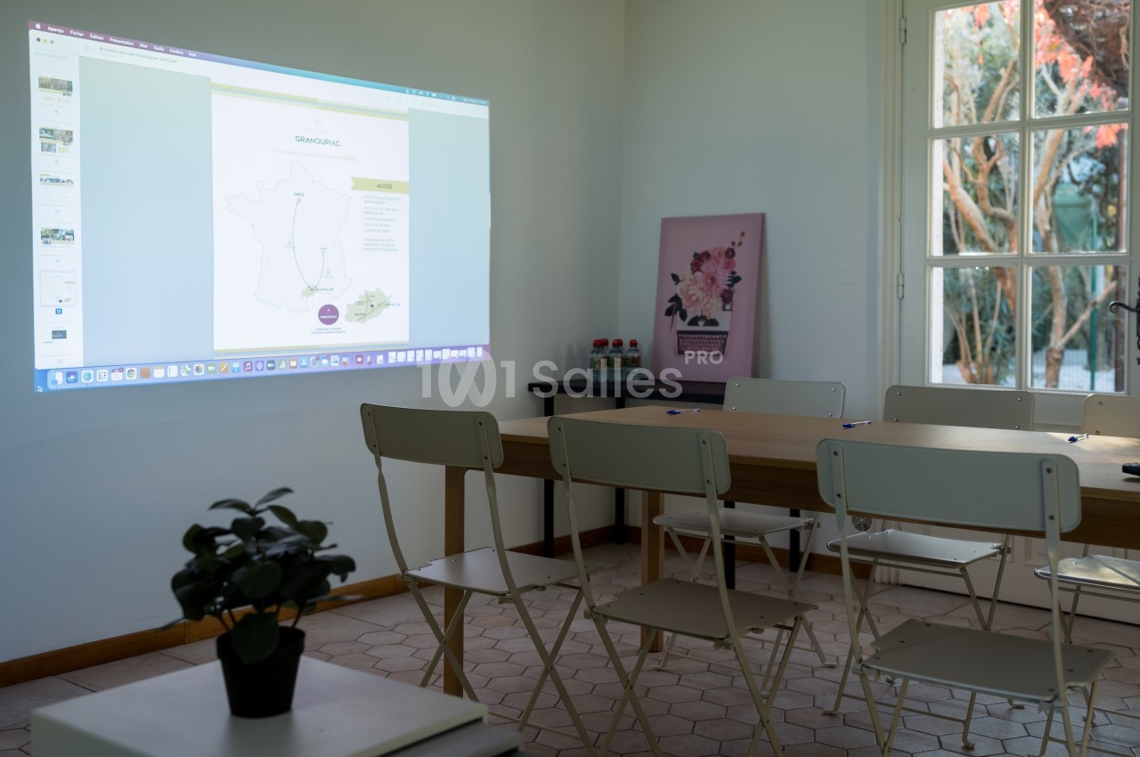 Salle de réunion lumineuse avec des chaises blanches, une table en bois et une présentation projetée au mur.