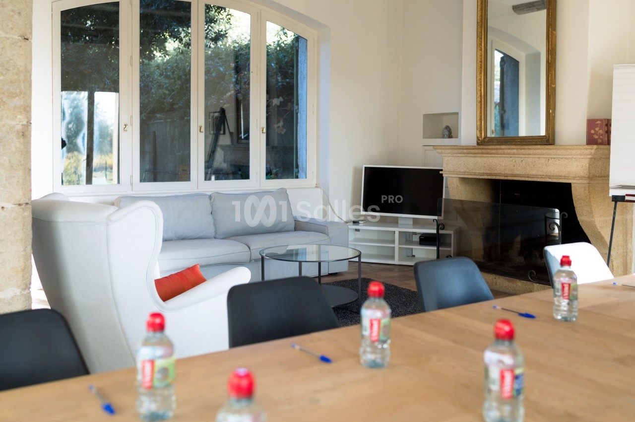 Salle lumineuse avec table en bois, chaises noires, canapé, cheminée, télévision et grandes fenêtres donnant sur un jardin.