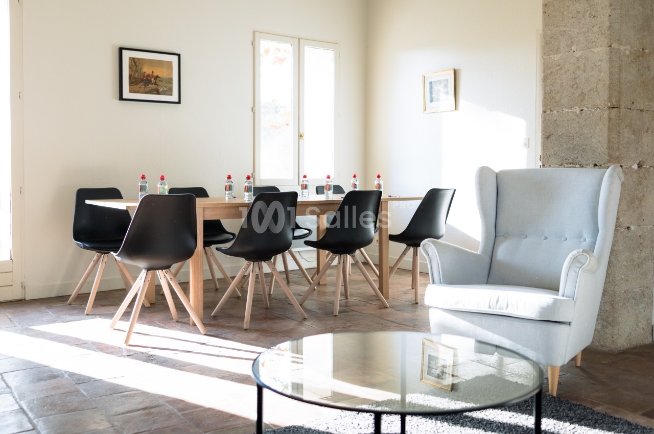 Salle lumineuse avec table en bois entourée de chaises noires, fauteuil gris et table basse en verre.