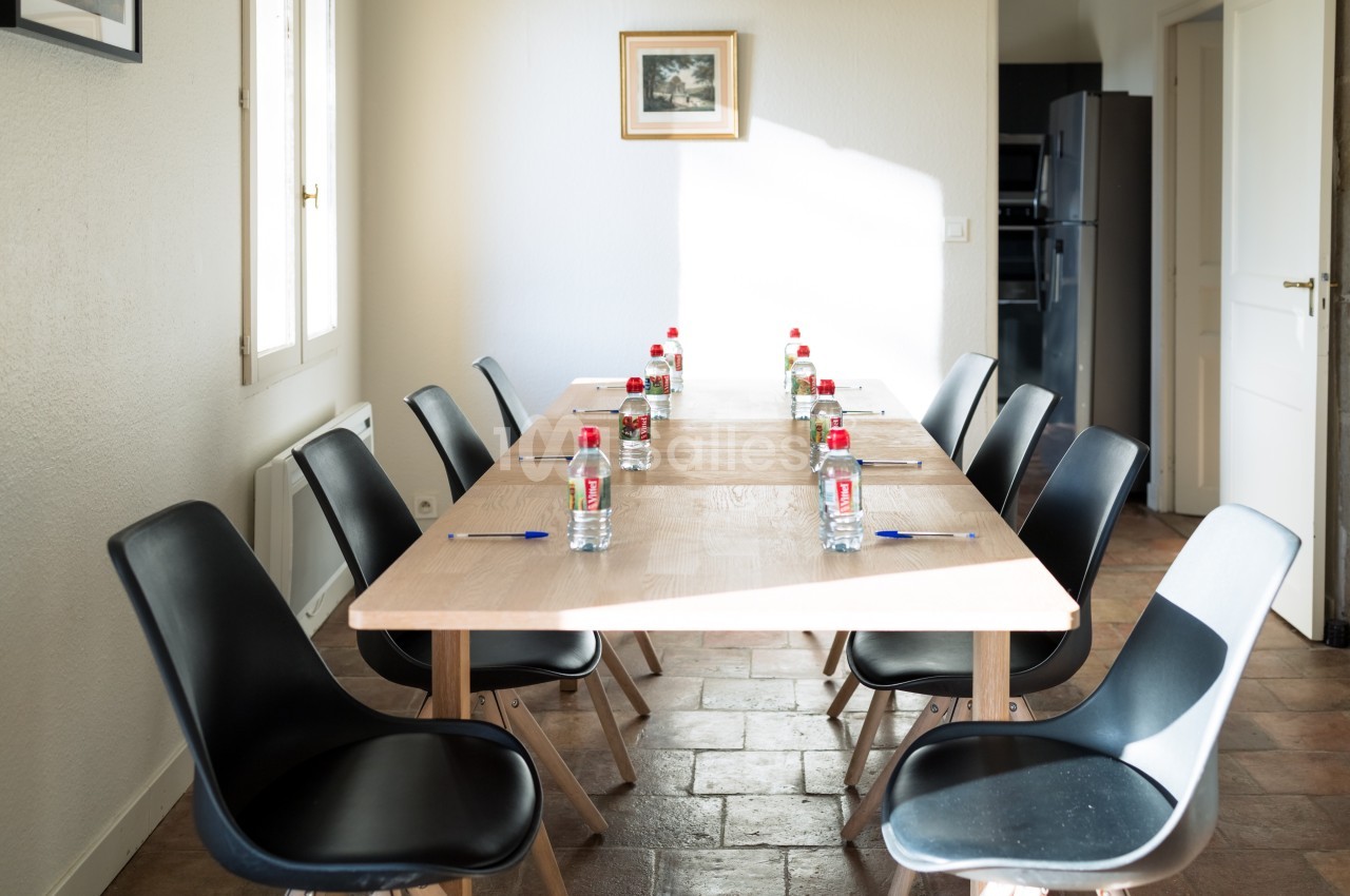 Table rectangulaire avec huit chaises noires, bouteilles d'eau et stylos disposés pour une réunion dans une pièce lumineuse.