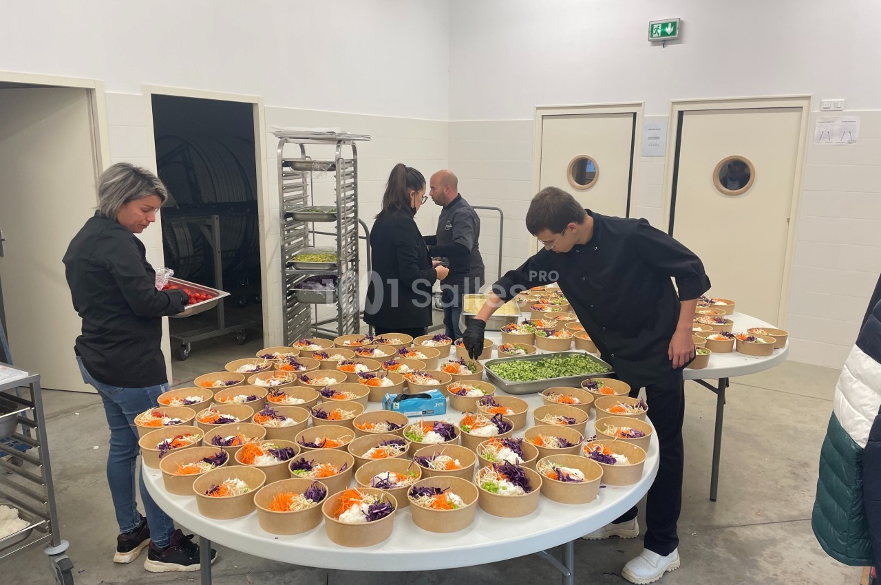 Des personnes préparent des salades colorées dans des bols en carton sur des tables dans une cuisine professionnelle.