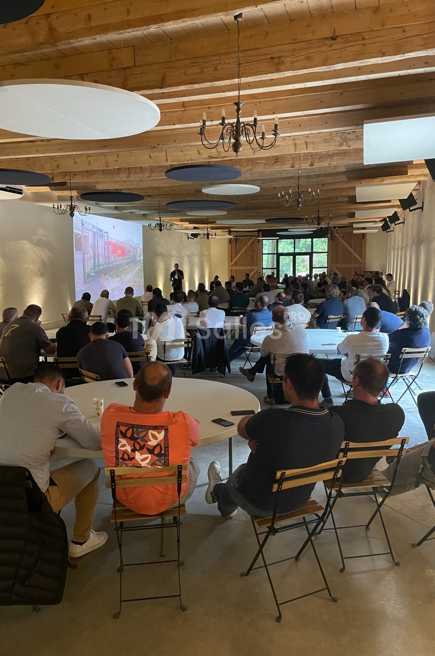 Des personnes assistent à une présentation projetée dans une salle avec un plafond en bois et des tables rondes.