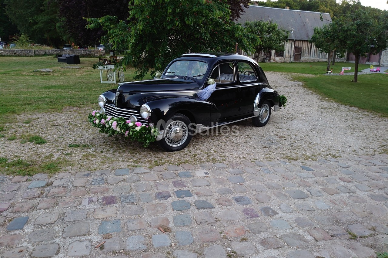 Voiture ancienne noire décorée de fleurs, stationnée sur une allée pavée dans un cadre champêtre.
