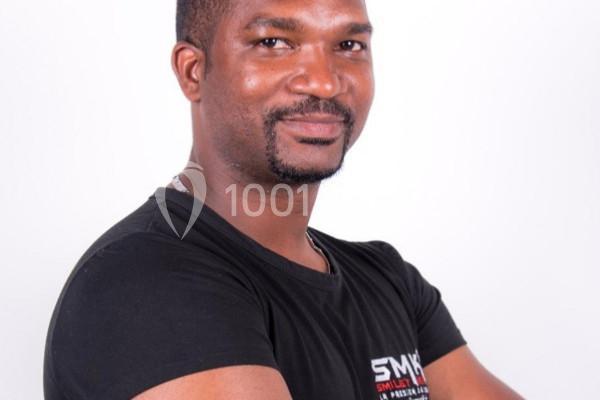Homme souriant portant un t-shirt noir, bras croisés, posé devant un fond blanc.