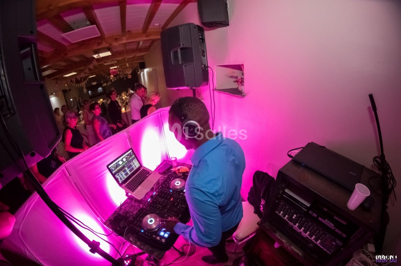 Un DJ mixe de la musique sur une console dans une salle éclairée par des lumières roses, avec des invités en arrière-plan.