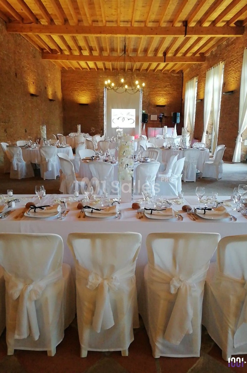Salle de réception avec tables dressées, nappes blanches, chaises décorées et éclairage naturel à travers de grandes…