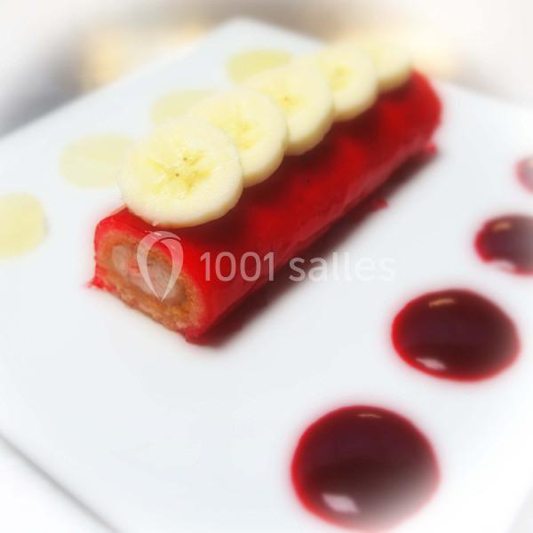 Dessert roulé recouvert de glaçage rouge, garni de rondelles de banane, avec des touches de coulis sur l'assiette blanche.