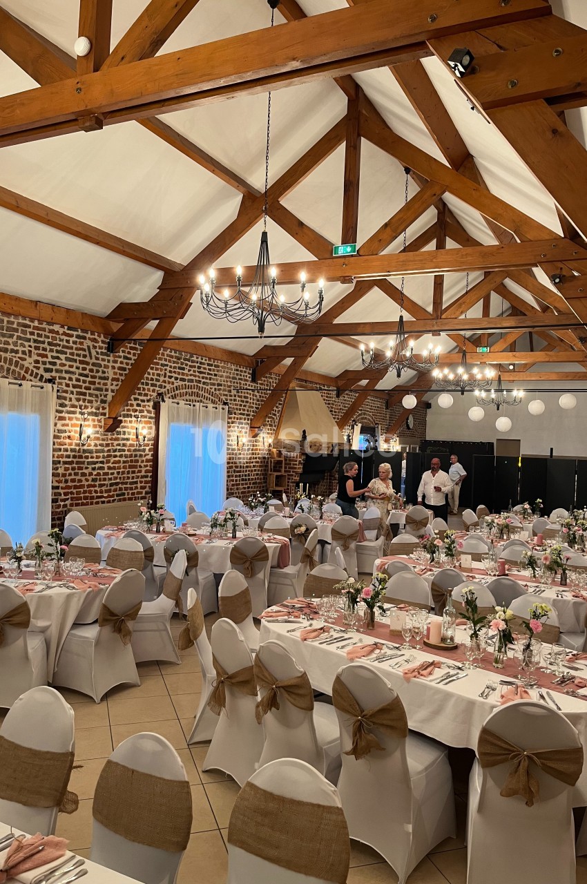Salle de réception décorée avec des tables dressées, nappes blanches, chaises ornées et poutres apparentes au plafond.