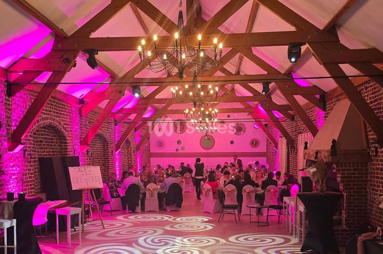 Salle de réception en briques avec poutres apparentes, éclairée par des lumières roses et des lustres, tables dressées.