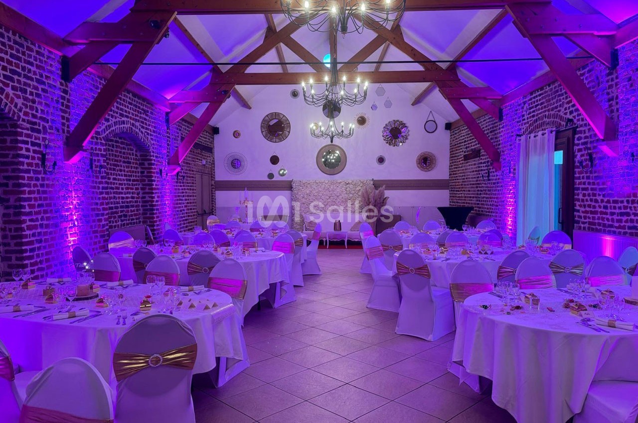 Salle de réception décorée avec des tables rondes dressées, éclairage violet et poutres apparentes au plafond.