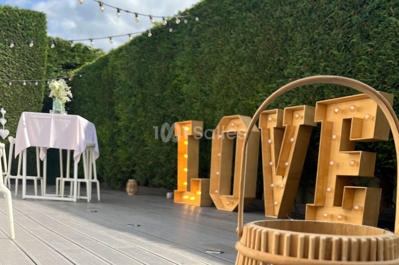 Décoration extérieure avec grandes lettres lumineuses ’LOVE’, table haute blanche et guirlandes suspendues, devant une haie.