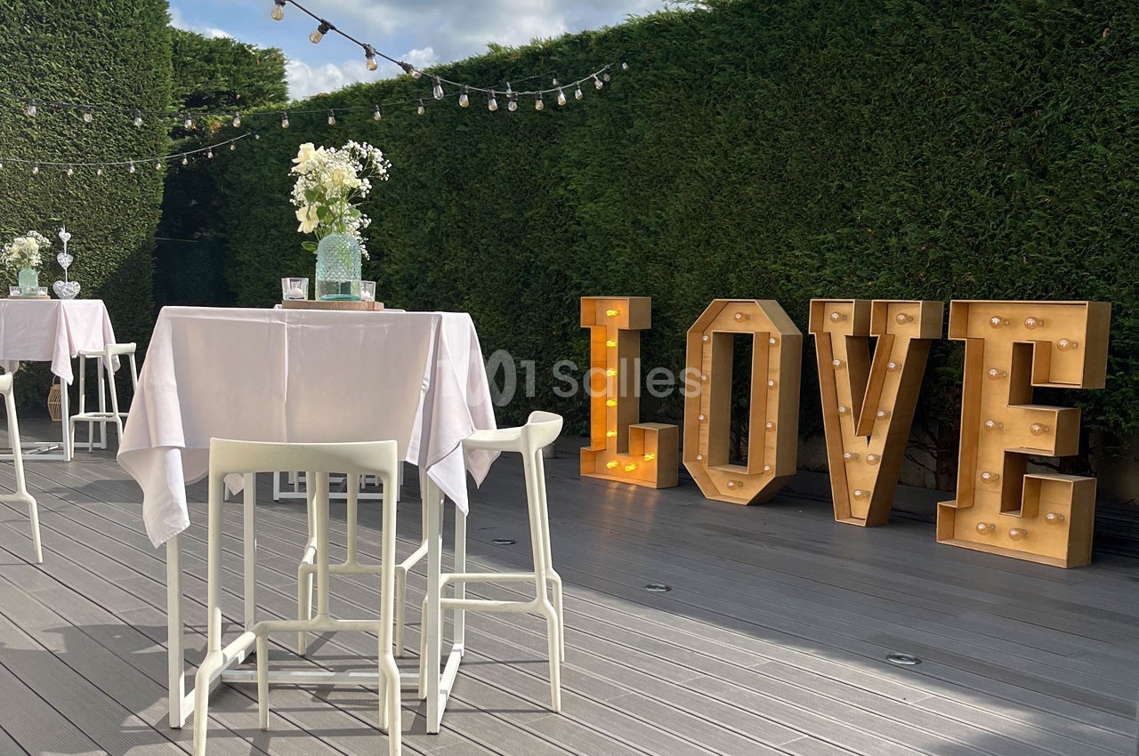 Terrasse décorée avec des tables hautes, guirlandes lumineuses et lettres géantes ’LOVE’ éclairées devant une haie.