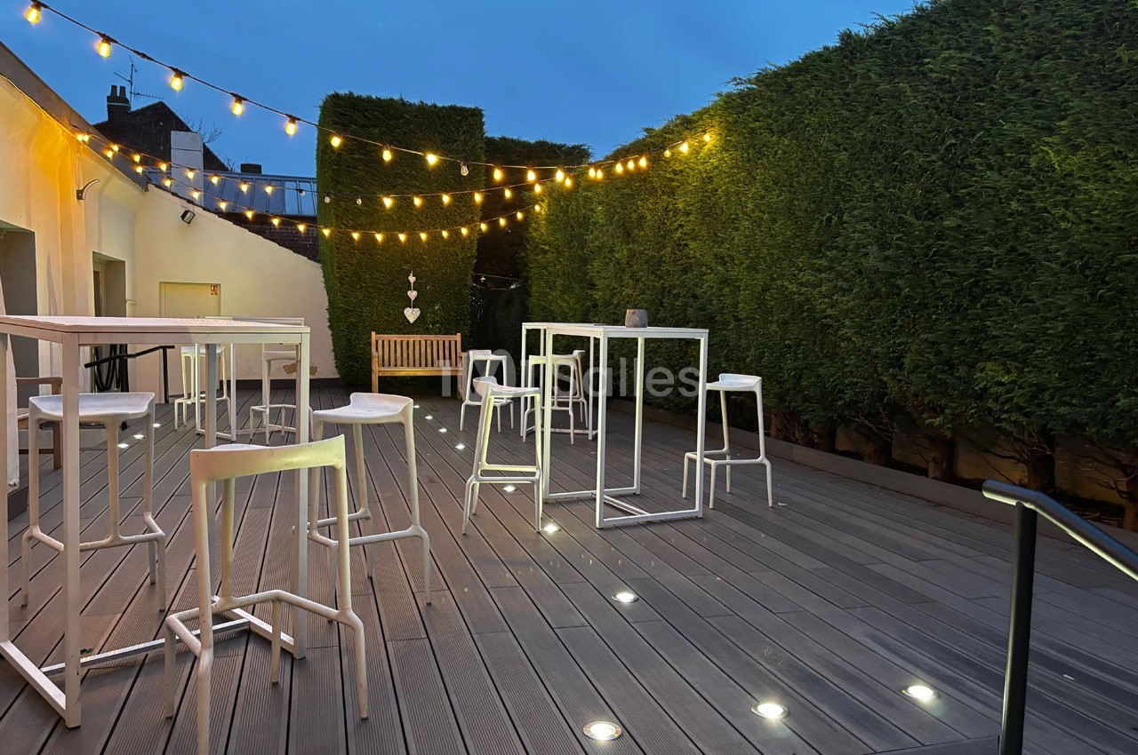 Terrasse en bois avec tables hautes, tabourets blancs, guirlandes lumineuses et haies taillées en soirée.