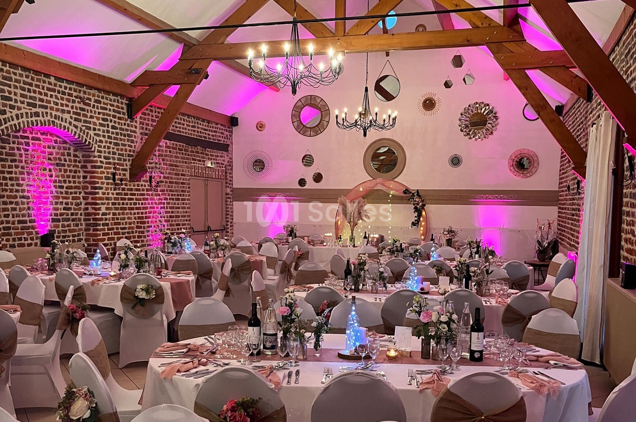 Salle de réception décorée avec des tables dressées, chaises ornées de nœuds, éclairage rose et murs en briques.