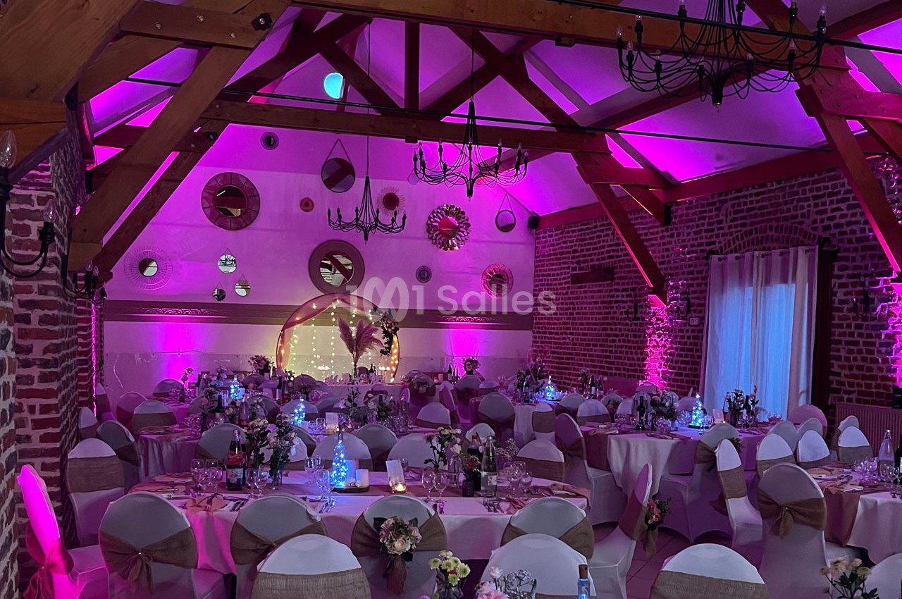Salle de réception décorée avec des tables rondes, des chaises habillées et un éclairage rose chaleureux.