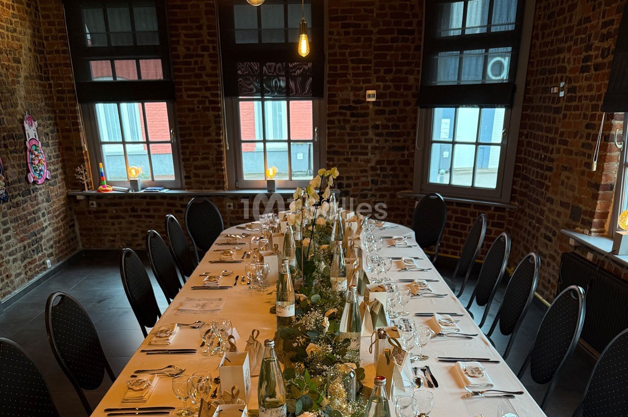 Table élégamment dressée pour un repas, décorée de fleurs et de bouteilles, dans une salle en briques avec grandes fenêtres.