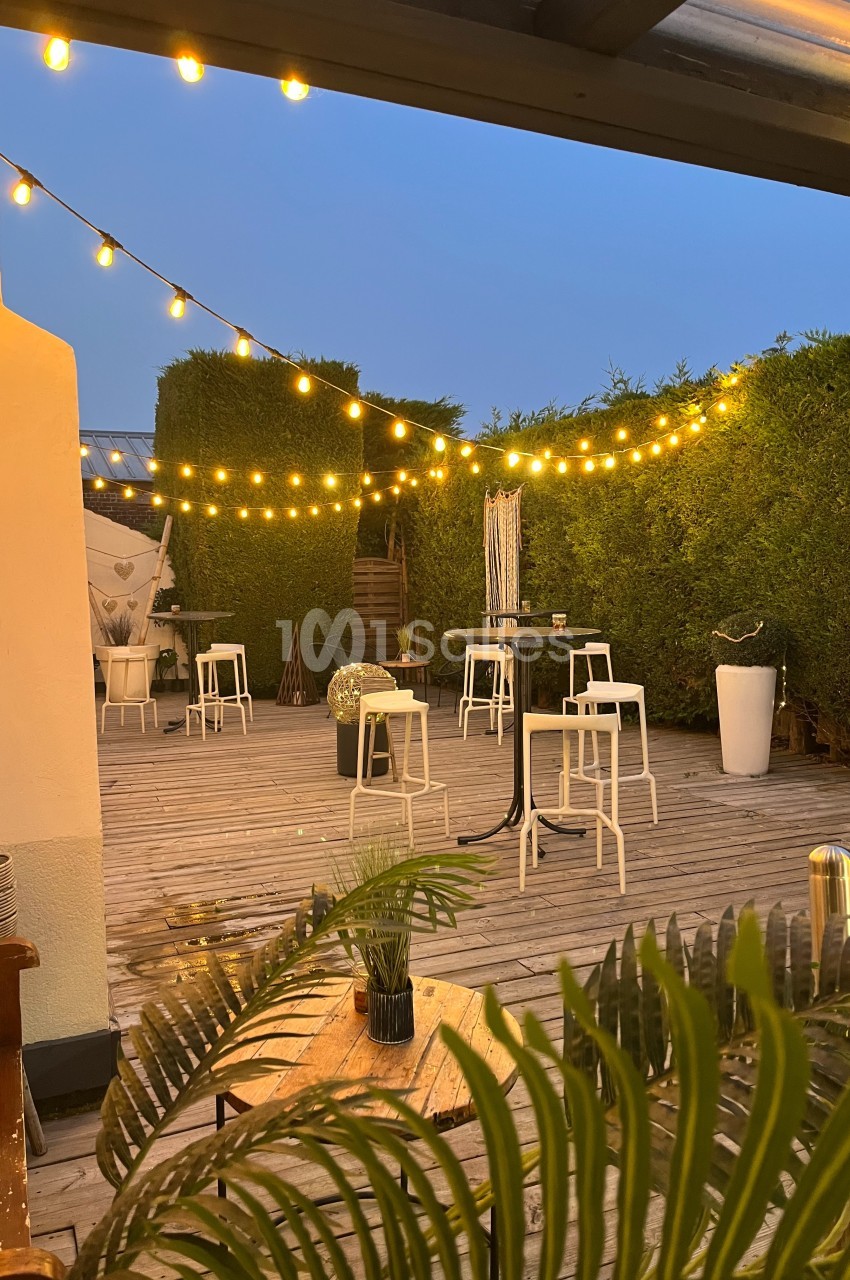 Terrasse en bois avec des guirlandes lumineuses suspendues, entourée de haies et aménagée avec tables et chaises blanches.